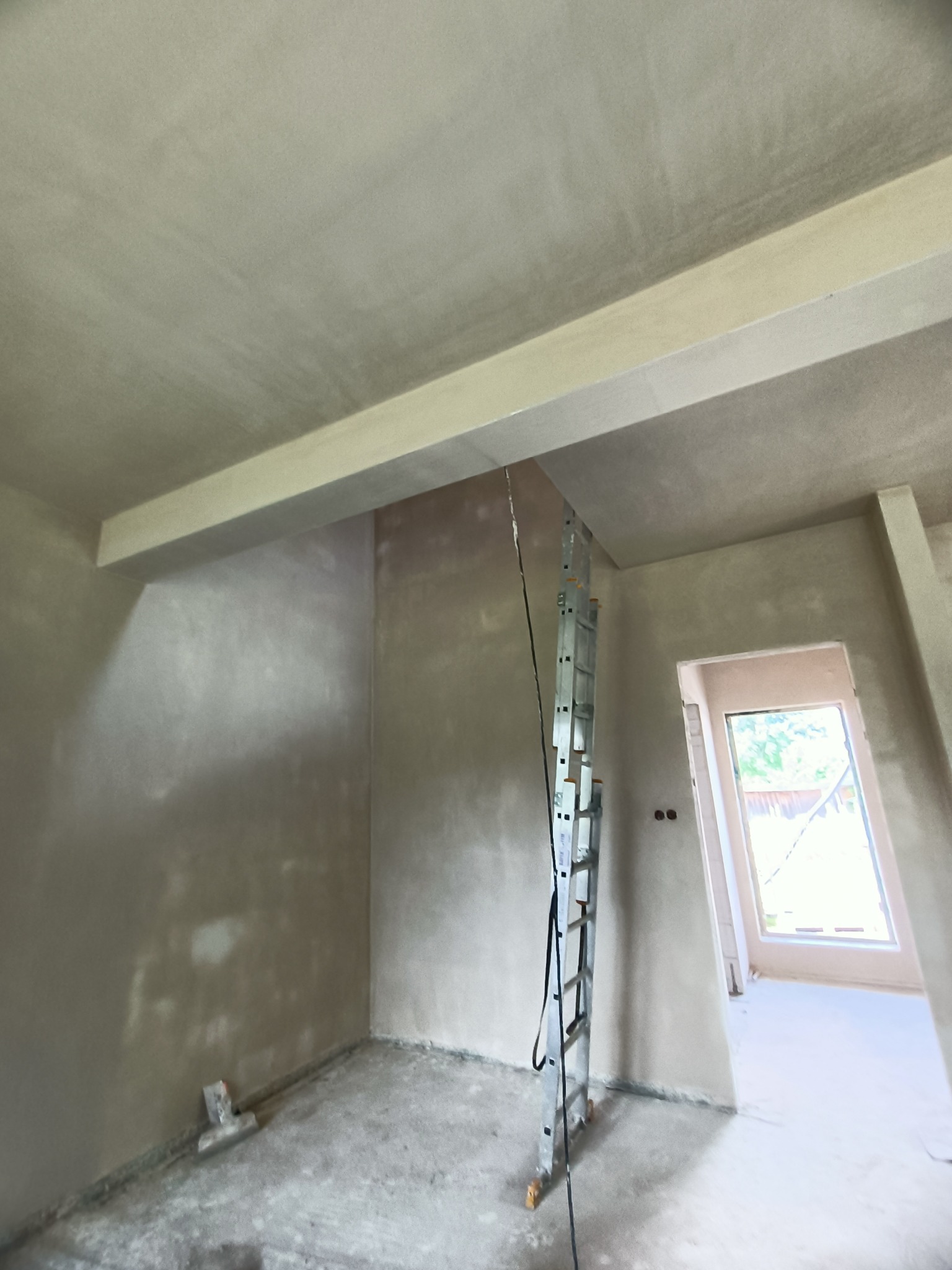 Wnętrze po nałożeniu świeżych tynków, widoczna aluminiowa drabina oparta o ścianę, narzędzie do wygładzania tynku leżące przy podłodze w rogu pomieszczenia, surowe oświetlenie.