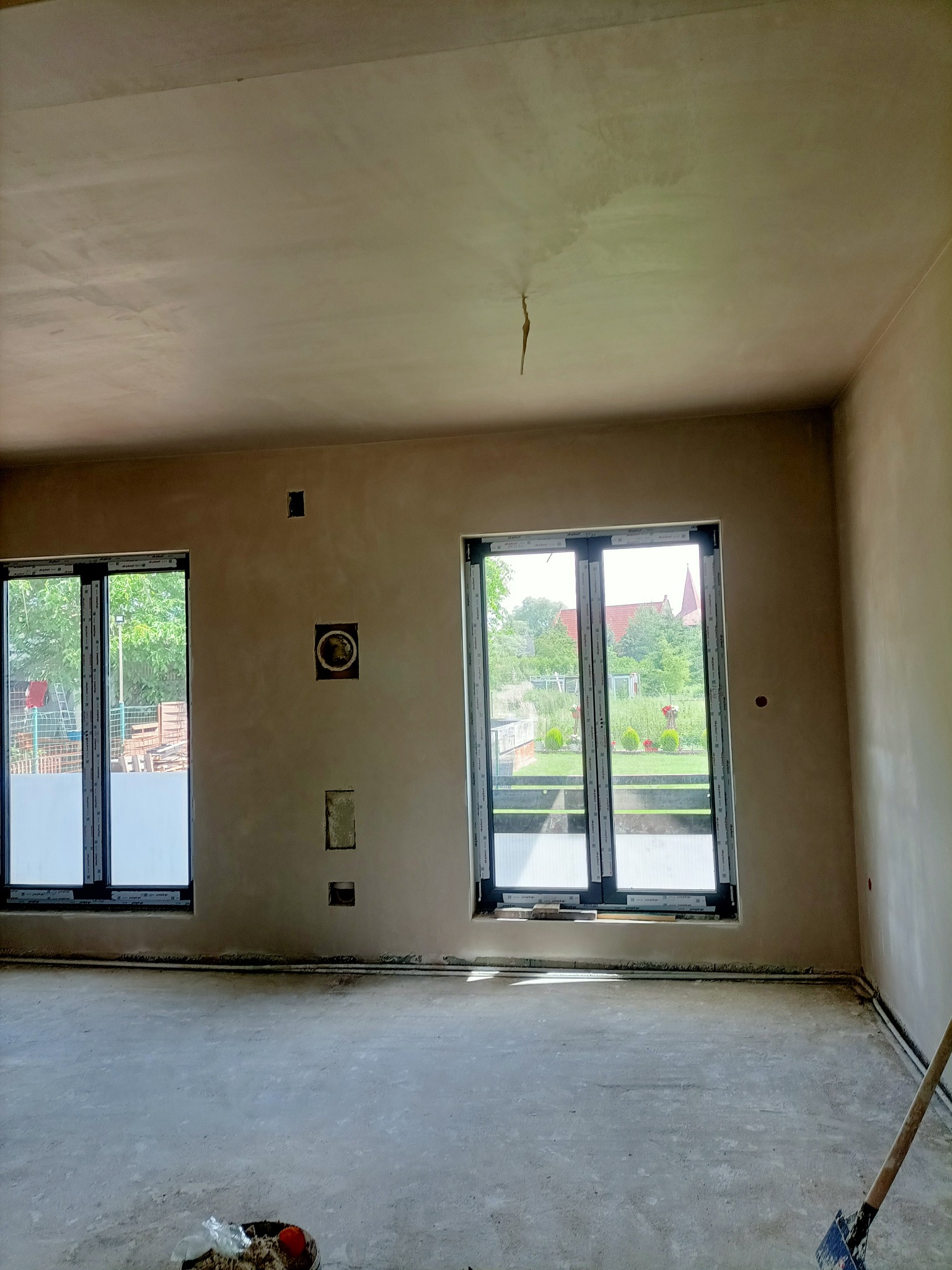 Wnętrze po nałożeniu gładzi gipsowej, widoczne okna z widokiem na zewnątrz, z pozostawionymi otworami instalacyjnymi w ścianie i niedoczyszczoną podłogą.