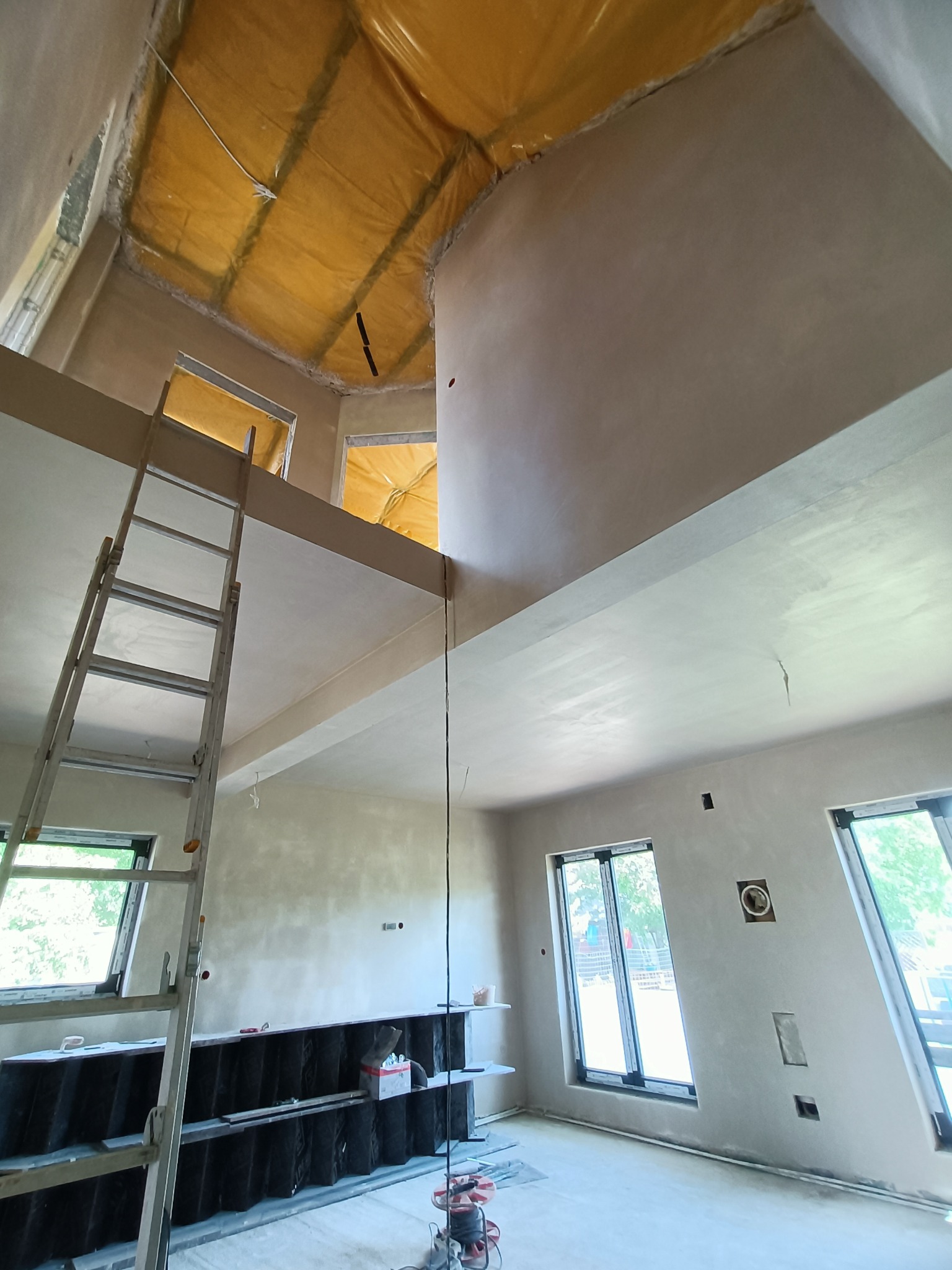Wnętrze domu w trakcie prac wykończeniowych, widok na wysoki sufit z nałożonymi świeżymi tynkami, zabezpieczona folią przestrzeń pod dachem, aluminiowa drabina oparta o ścianę, widoczne okna...