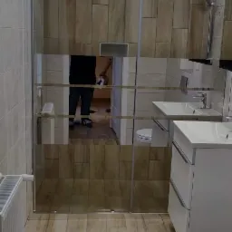 Nowoczesna łazienka z kabiną prysznicową ze szklanymi drzwiami przesuwnymi, umywalką w białej szafce i płytkami imitującymi drewno na ścianach i podłodze. W lustrze odbija się osoba wykonująca...