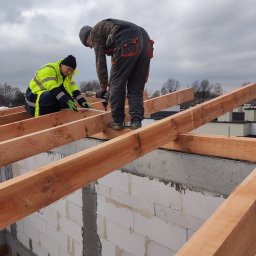 Global Labs sp. z o.o. - Dwóch dekarzy montuje drewnianą konstrukcję dachu na murowanym budynku. Jeden mierzy, drugi przykręca. Widok z dołu, pochmurne niebo w tle.