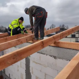 Dwóch dekarzy montuje drewnianą konstrukcję dachu na murowanym budynku. Jeden mierzy, drugi przykręca. Widok z dołu, pochmurne niebo w tle.
