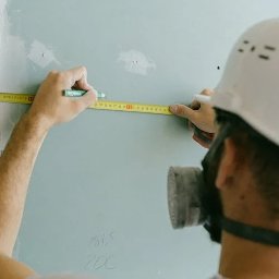 Global Labs sp. z o.o. - Pracownik w masce i kasku mierzy ścianę miarką, zaznaczając wymiary ołówkiem. Ściana jest częściowo wykończona, widoczne ślady po łataniu.