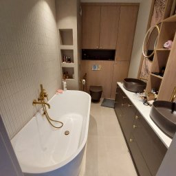 Global Labs sp. z o.o. - Nowoczesna łazienka z wolnostojącą wanną, złotą armaturą i umywalkami nablatowymi w odcieniach brązu. Minimalistyczne szafki i półki w zabudowie.