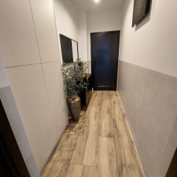 Global Labs sp. z o.o. - Nowoczesny przedpokój z drewnopodobną podłogą, szarą lamperią i minimalistyczną szafą w zabudowie. Dekoracyjna roślina w wazonie dodaje wnętrzu charakteru.