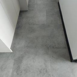 Global Labs sp. z o.o. - Szara, matowa podłoga z płytek imitujących kamień, ułożona w korytarzu. Widoczne białe szafki po lewej i listwa przypodłogowa po prawej stronie.