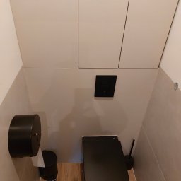 Global Labs sp. z o.o. - Nowoczesna toaleta w odcieniach beżu i czerni. Podwieszana muszla klozetowa, drewnopodobna podłoga, minimalistyczne szafki nad stelażem. Uwagę zwraca czarny dozownik papieru.