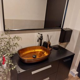 Global Labs sp. z o.o. - Nowoczesna łazienka z umywalką w odcieniach złota, czarną armaturą i szafką z szufladami. Minimalistyczne lustro i dekoracyjne gałązki dodają elegancji.