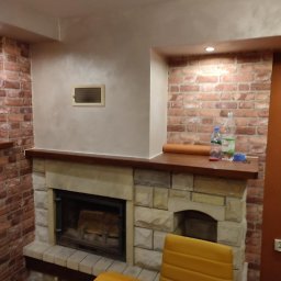 Global Labs sp. z o.o. - Kamienny kominek z drewnianym blatem, otoczony ścianą z cegły i gładzią. Na blacie butelki z wodą i zwinięty materiał. Część stołu i krzesła w kadrze.