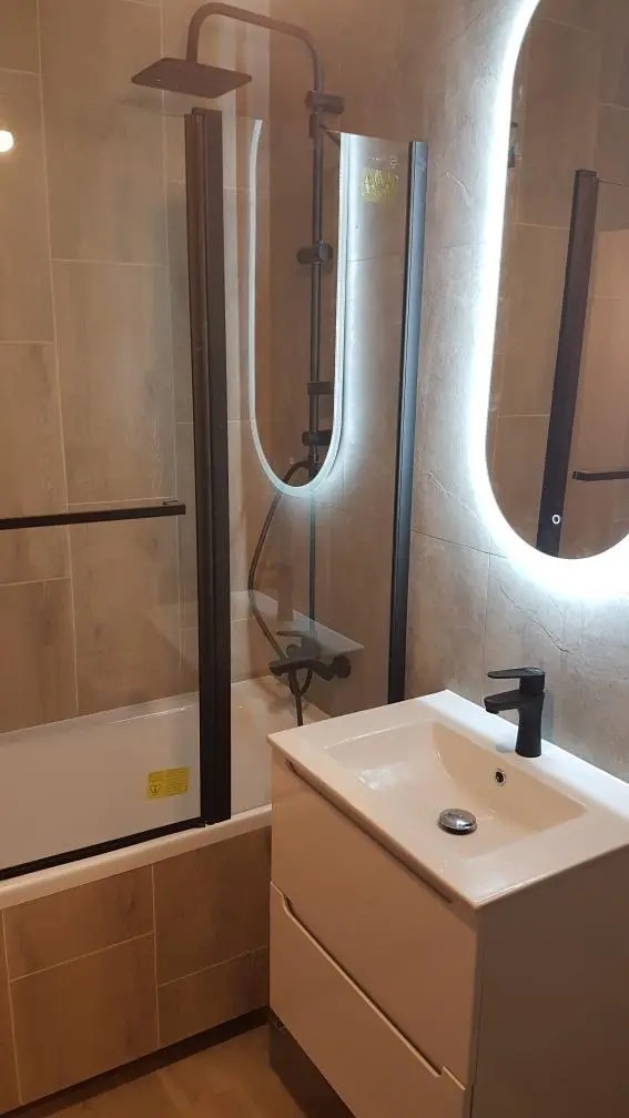 Nowoczesna łazienka z beżowymi płytkami, kabiną prysznicową z czarnymi ramami, białą szafką z umywalką i podświetlanym lustrem owalnym.