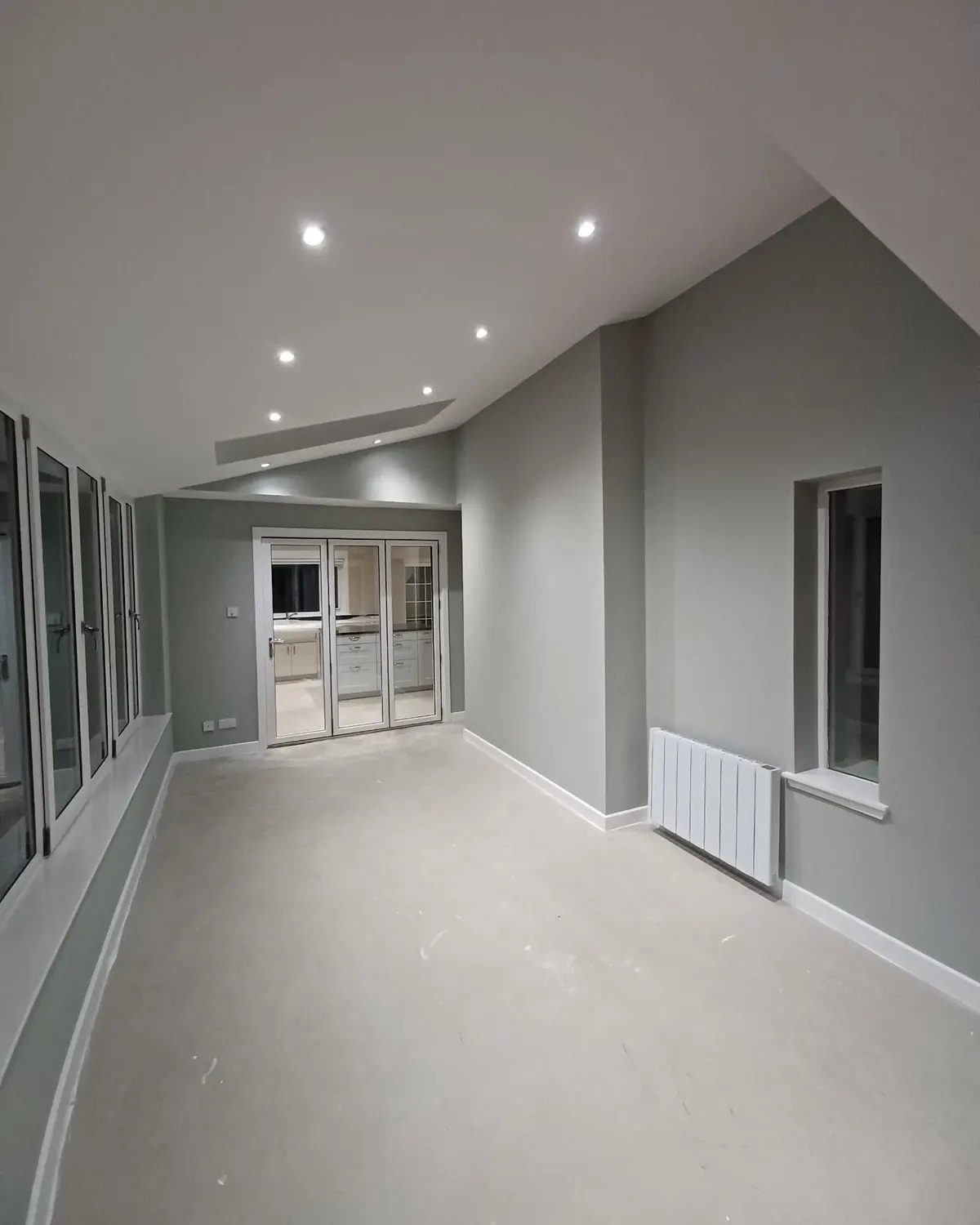 Jasne wnętrze po malowaniu: szare ściany, białe listwy przypodłogowe i sufit z oświetleniem punktowym. Widoczne okna i grzejnik. Minimalistyczny design.