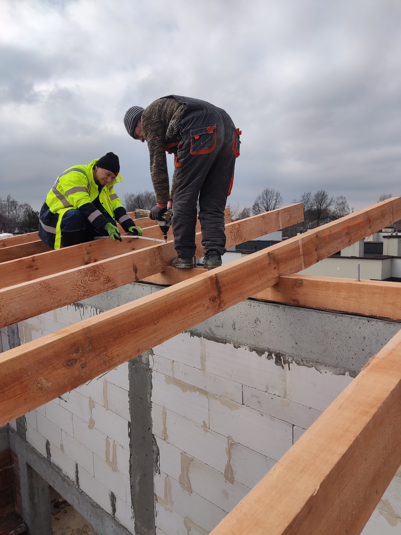 Dwóch dekarzy montuje drewnianą konstrukcję dachu na murowanym budynku. Jeden mierzy, drugi przykręca. Widok z dołu, pochmurne niebo w tle.