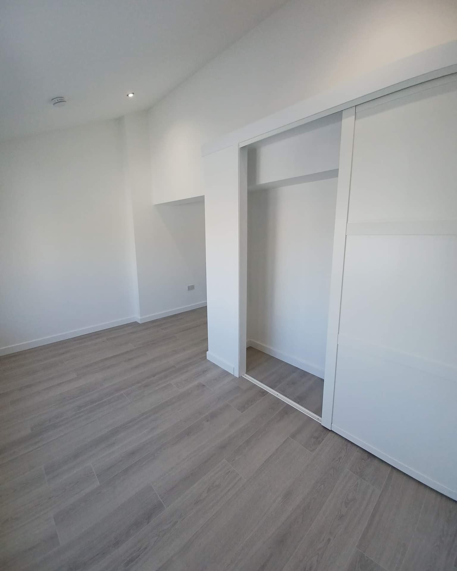 Jasne wnętrze pokoju z szarymi panelami podłogowymi, białymi ścianami i wnękową szafą z przesuwnymi drzwiami. Minimalistyczny styl wykończenia wnętrza.