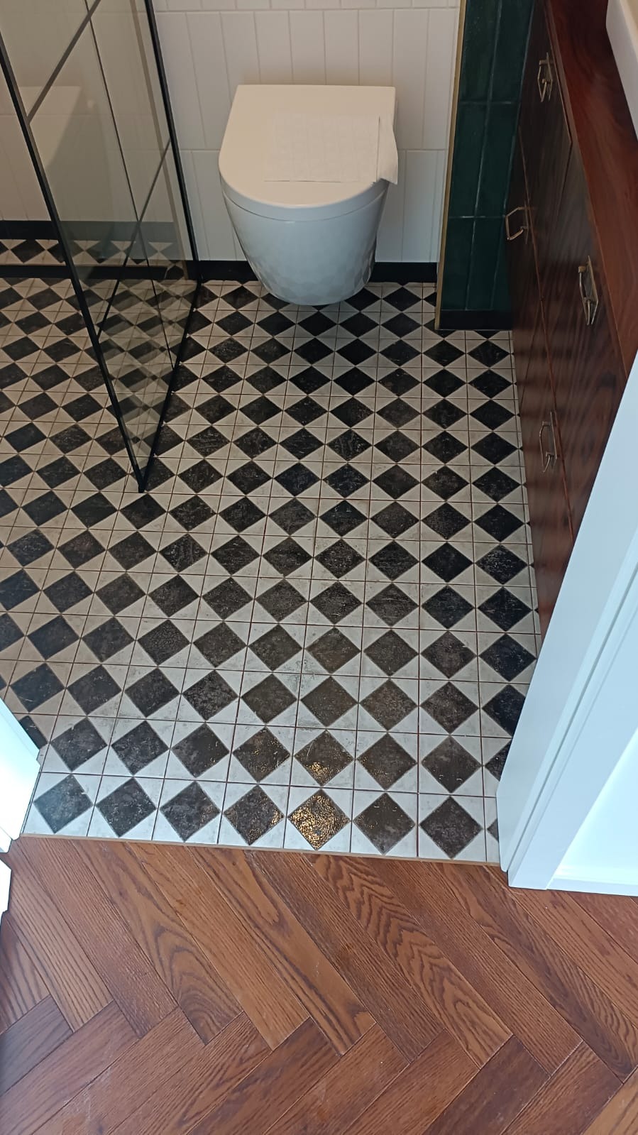 Wykończona łazienka z białą toaletą, kabiną prysznicową i wzorzystą podłogą w romby, przechodzącą w parkiet ułożony w jodełkę. Widok z góry, z otwartymi drzwiami.