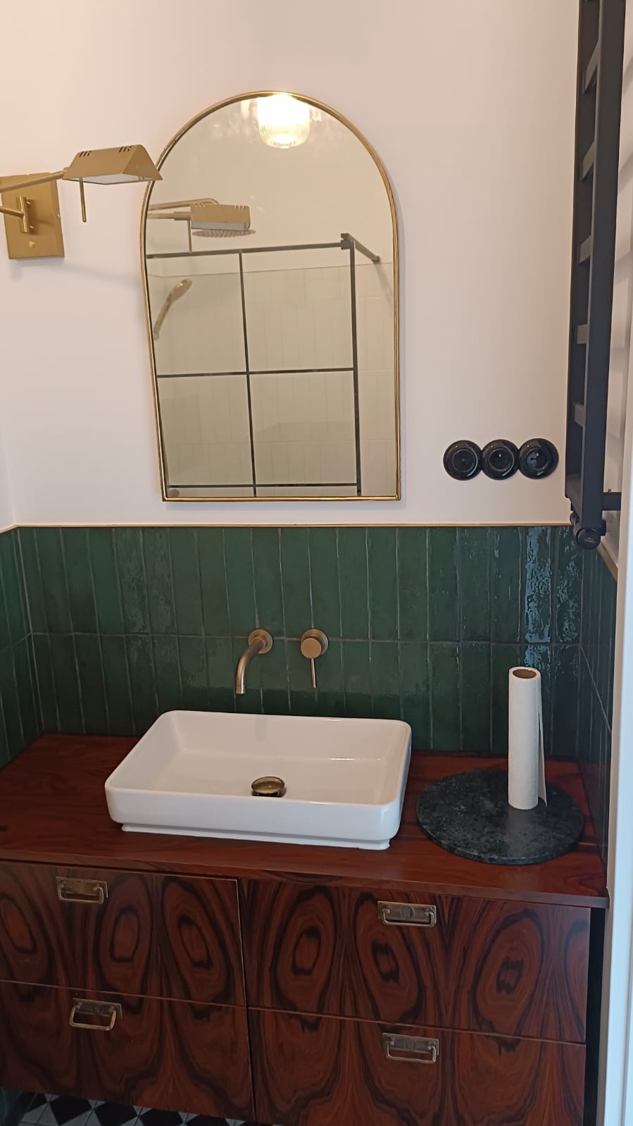 Wykończona łazienka w stylu retro: biała umywalka nablatowa na szafce z forniru, zielone płytki na ścianie, złota armatura i lustro w złotej ramie, czarny grzejnik.
