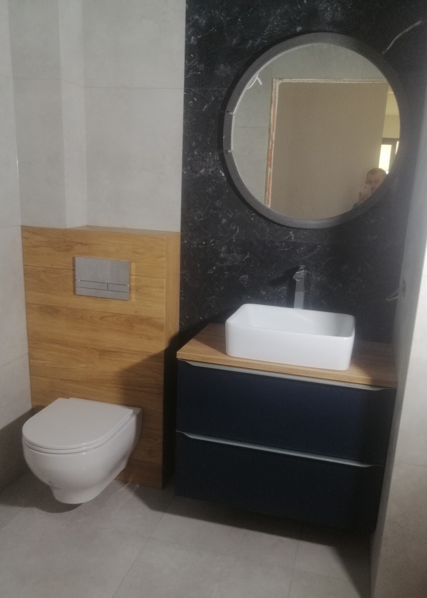 Wykończona łazienka: biała ceramika, umywalka na szafce z szufladami, okrągłe lustro na czarnej ścianie z marmurkowym wzorem, drewniane panele na ścianie.
