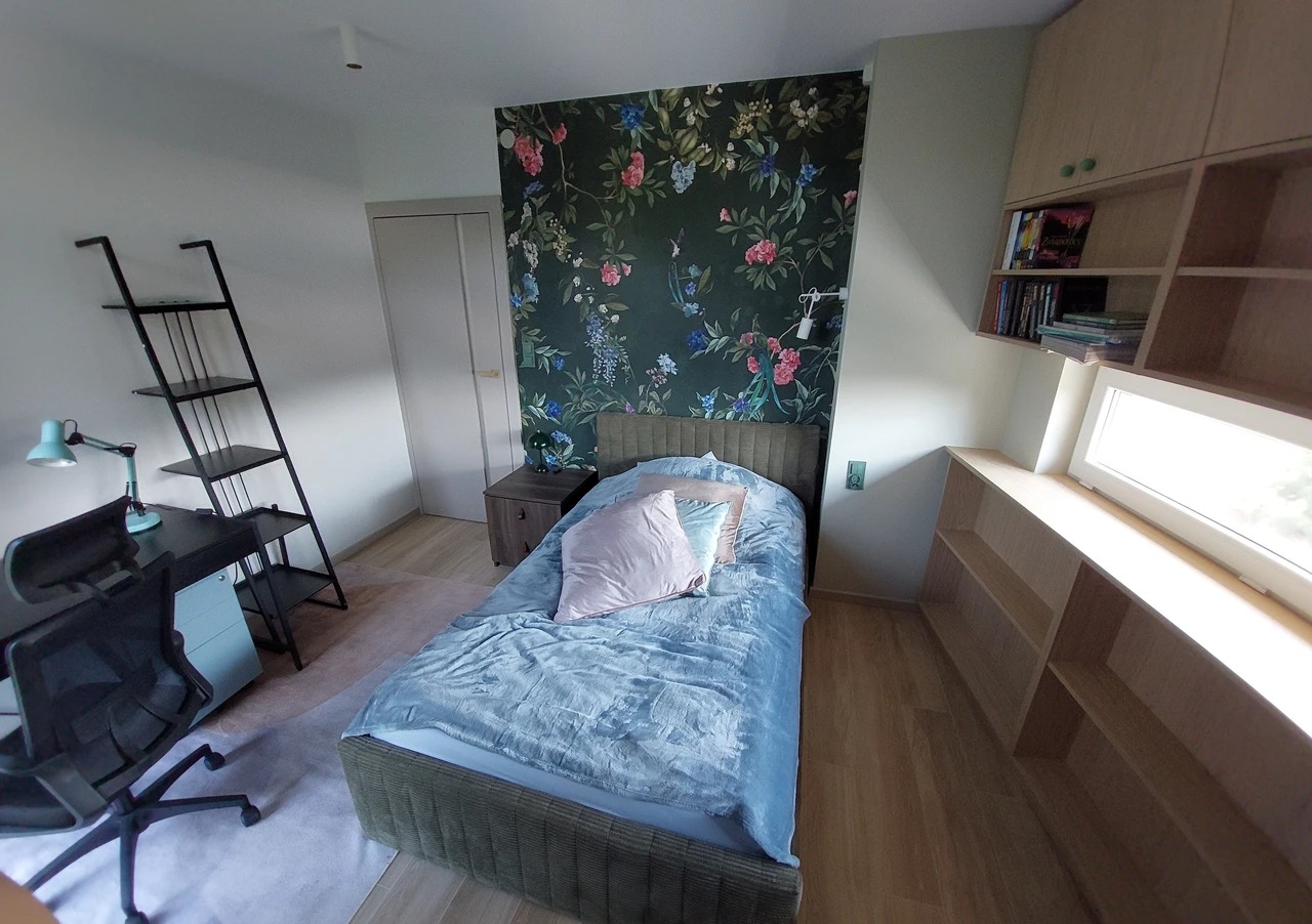 Pokój młodzieżowy po remoncie: łóżko z niebieską pościelą na tle ściany z motywem kwiatowym, biurko z lampką i półki pod oknem. Jasne drewno, nowoczesny design.