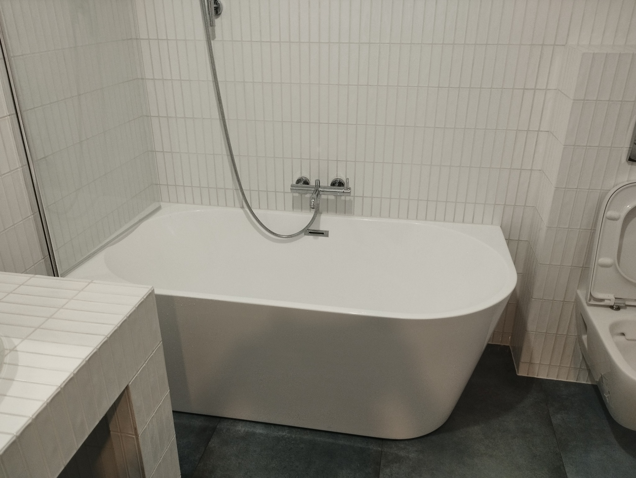 Nowoczesna, biała wanna wolnostojąca z chromowaną armaturą na tle ściany z białych, pionowych płytek w wyremontowanej łazience. Minimalistyczny design.