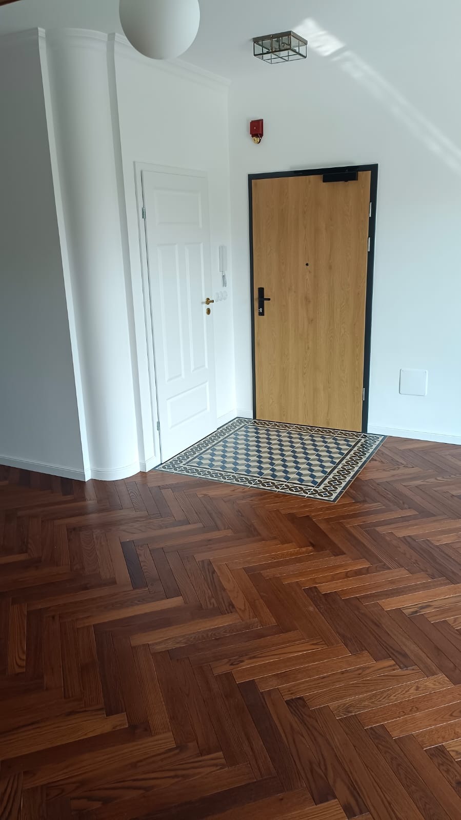 Parkiet w jodełkę, dywanik z geometrycznym wzorem przy drzwiach, białe ściany i drzwi. Nowoczesne wykończenie wnętrza z dbałością o detale.