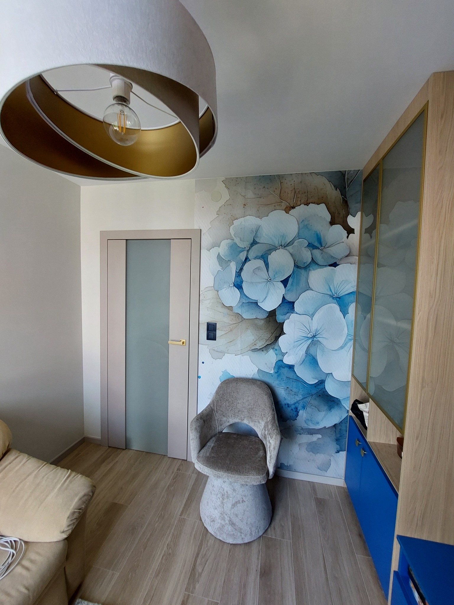 Stylowy pokój z fototapetą w kwiaty, designerską lampą i nowoczesną szafką z niebieskimi frontami. Widoczna także szara tapicerowana krzesło i drzwi z przeszkleniem.