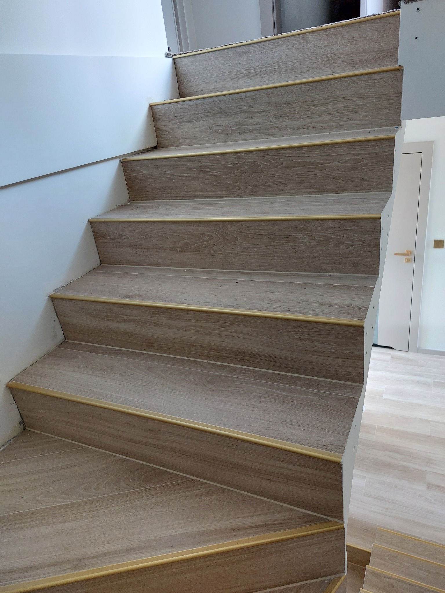 Wykończone schody z drewnopodobnymi panelami i złotymi listwami, widok z dołu. Na ścianie biała farba, w tle drzwi i podłoga z płytek.