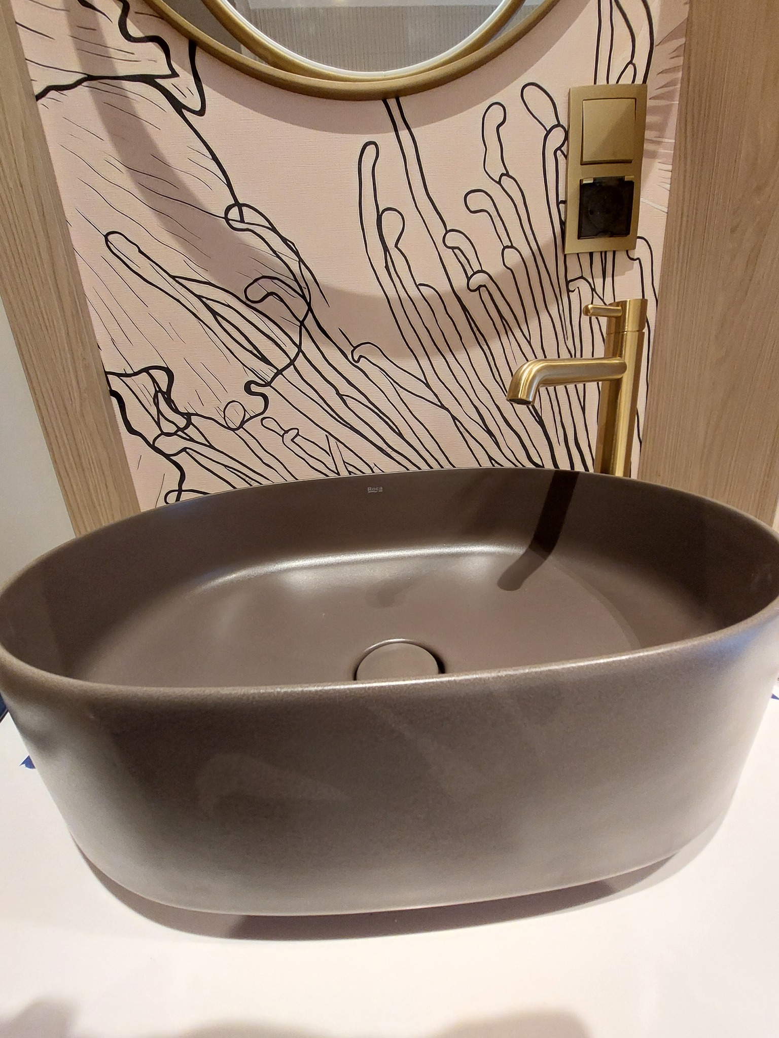 Stylowa, brązowa umywalka nablatowa Roca z złotą baterią, na tle wzorzystej tapety i okrągłego lustra. Detal łazienki po remoncie.