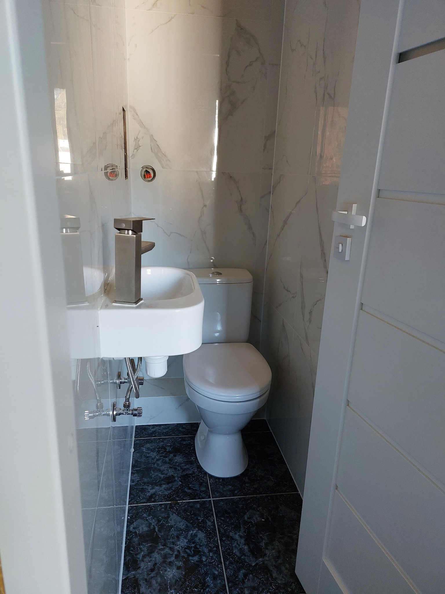 Nowoczesna, mała łazienka z marmurowymi płytkami, wiszącą umywalką i toaletą. Czarne płytki podłogowe z delikatnym wzorem. Białe drzwi wejściowe z widoczną klamką.