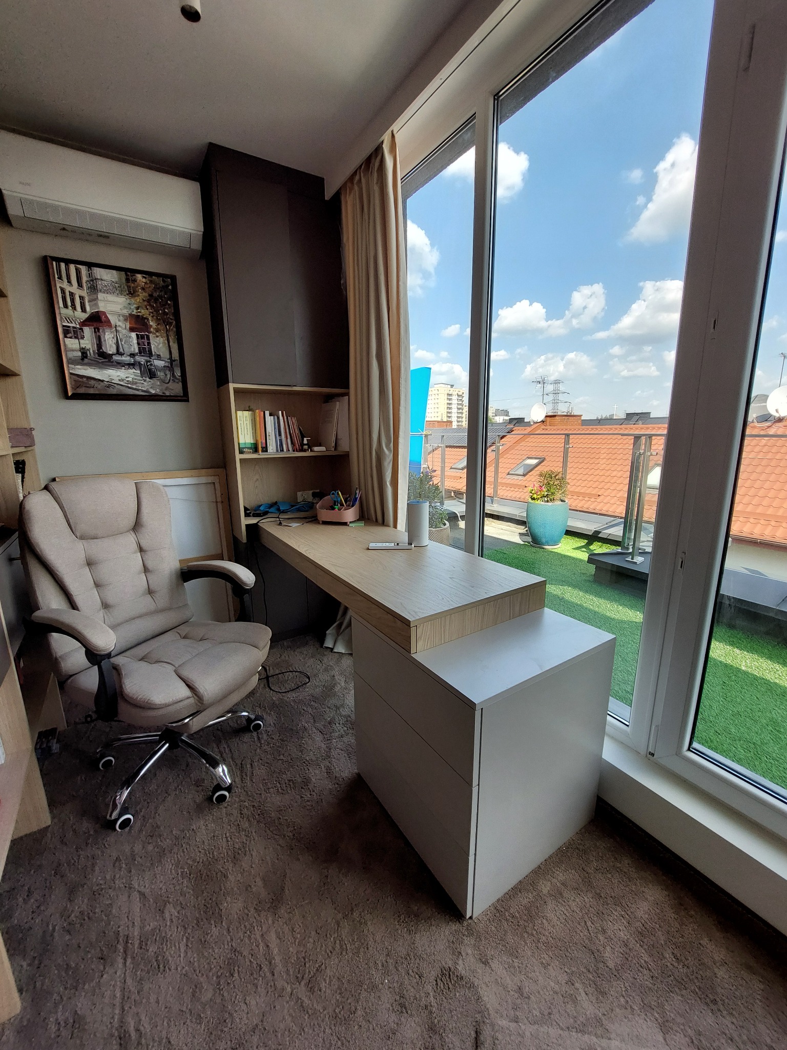 Nowoczesny pokój biurowy z dużym oknem, widok na dachy i sztuczną trawę. Jasne biurko z szufladami, ergonomiczne krzesło. Obraz na ścianie i klimatyzacja.