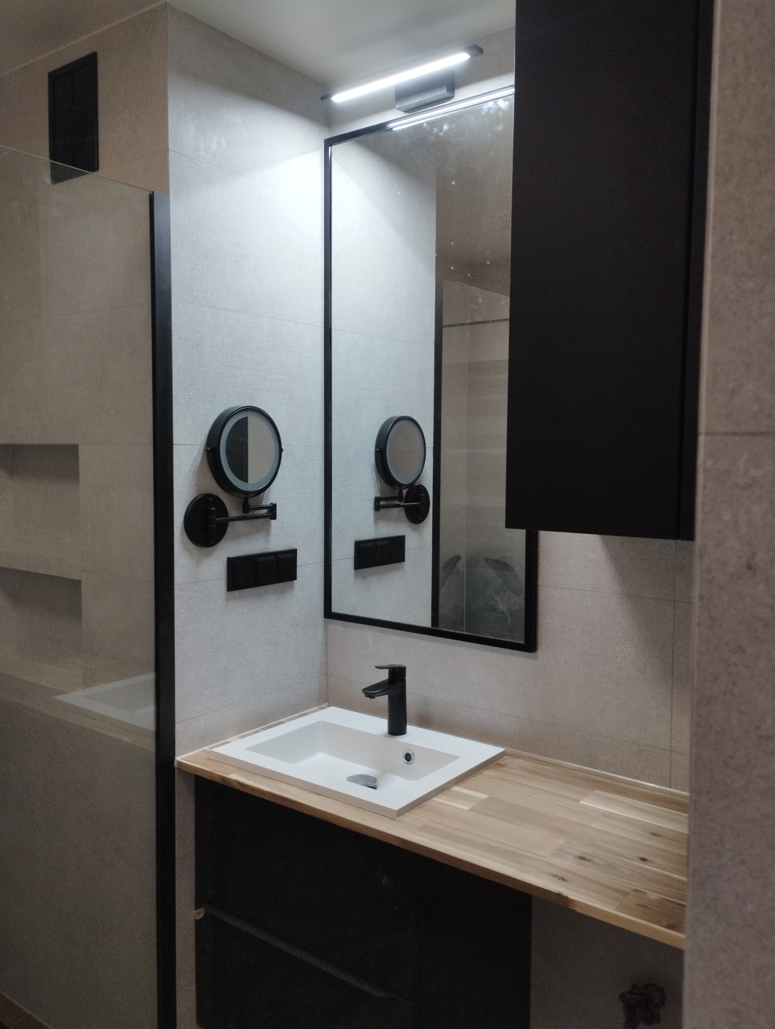 Nowoczesna łazienka z lustrem w czarnej ramie, umywalką nablatową i drewnianym blatem. Dwa podświetlane lusterka kosmetyczne na ścianie z szarych płytek.