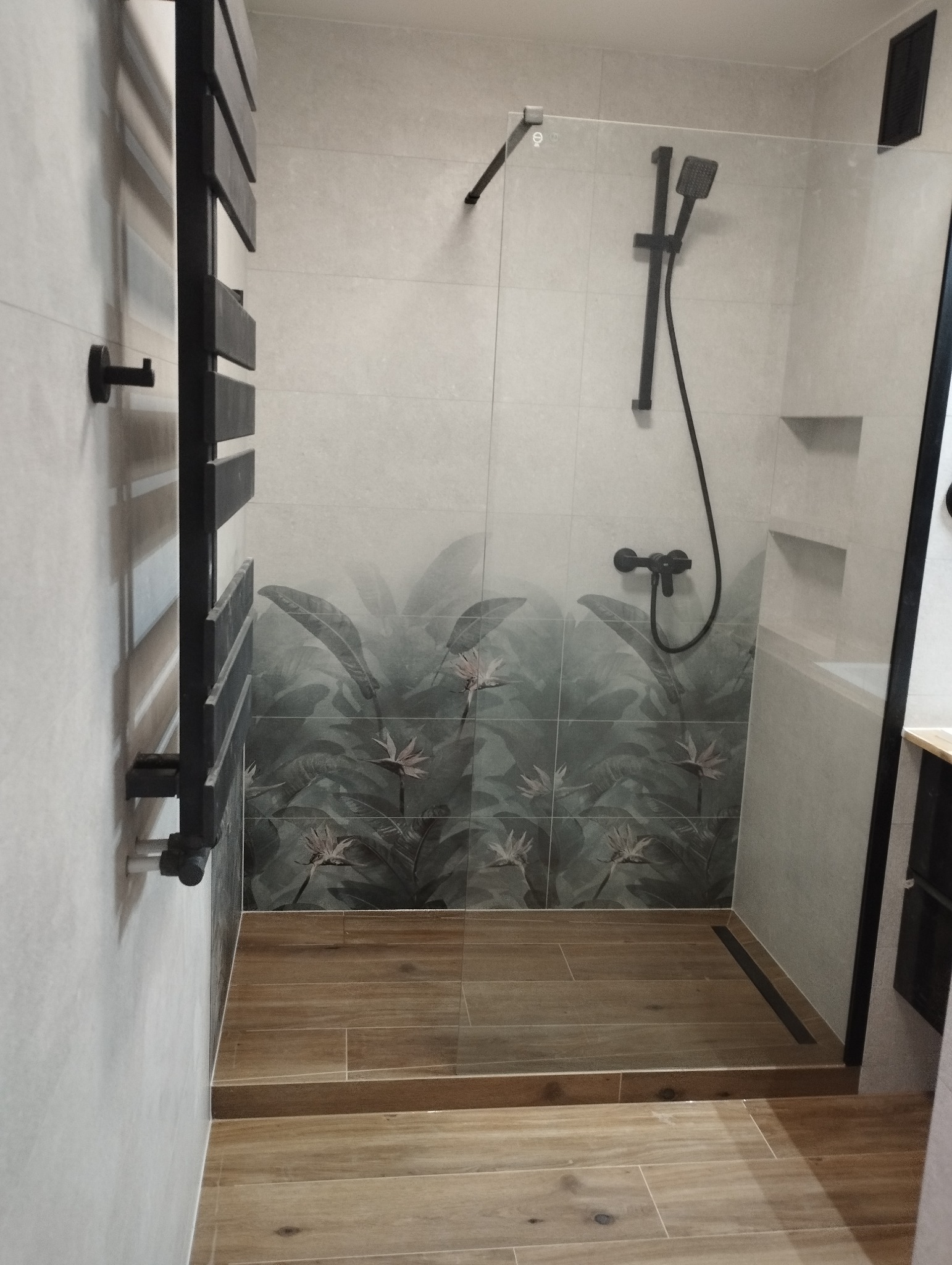 Nowoczesna kabina prysznicowa z motywem roślinnym na płytkach, czarna armatura i grzejnik drabinkowy. Drewnopodobne płytki na podłodze.