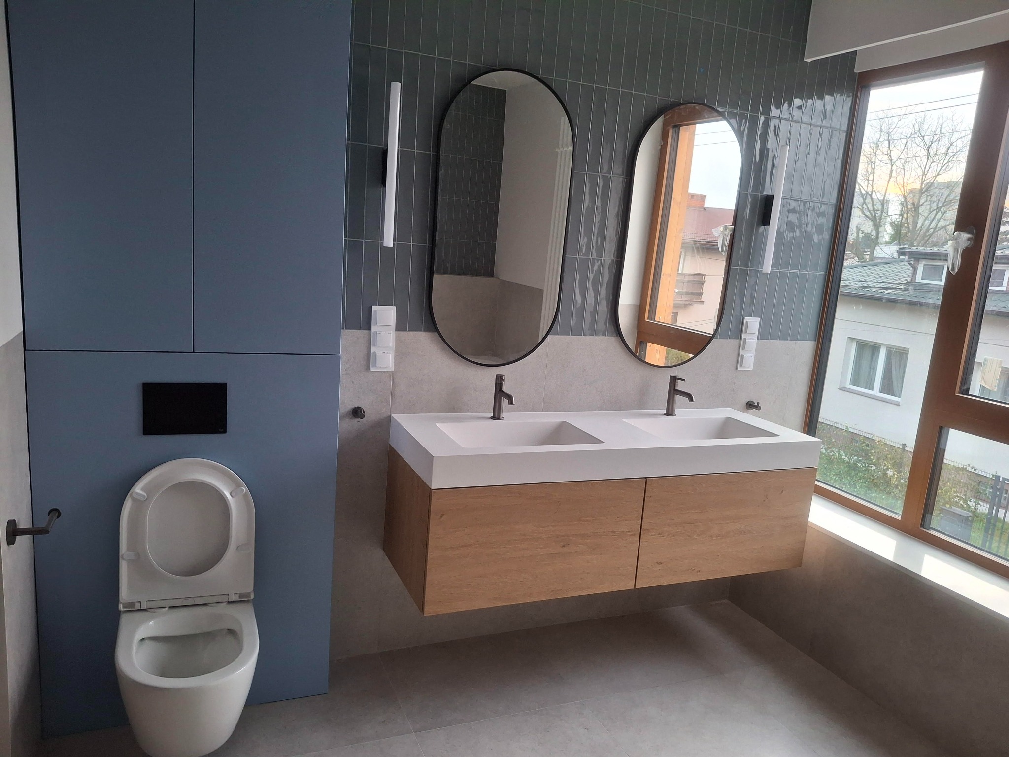 Nowoczesna łazienka z podwójną umywalką na drewnianej szafce, owalnymi lustrami i toaletą w zabudowie. Szaro-niebieska kolorystyka, pionowe płytki i duże okno.
