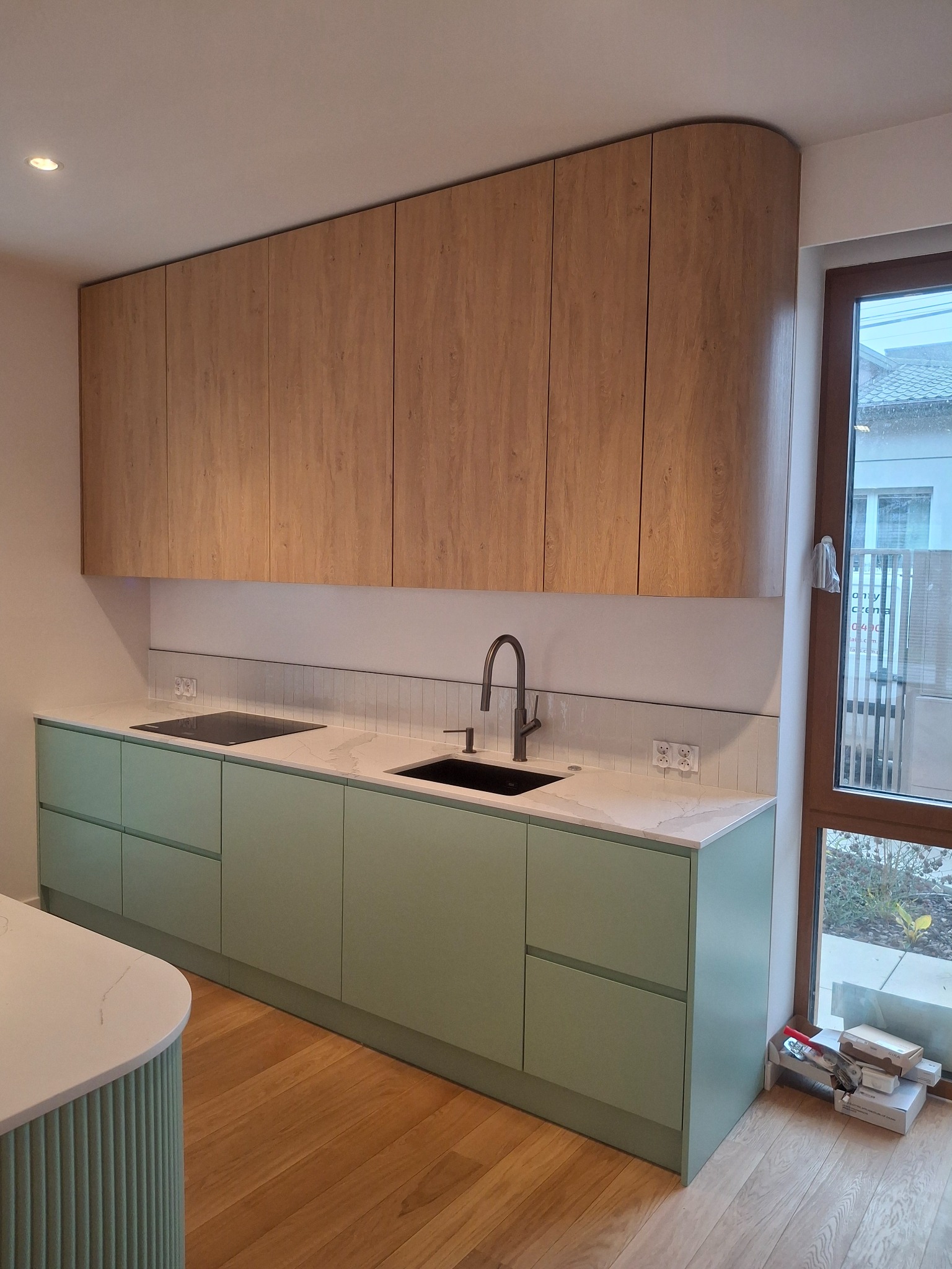 Nowoczesna kuchnia w pastelowych odcieniach zieleni i drewna. Zabudowa kuchenna z blatem imitującym marmur, czarna armatura i płyta indukcyjna. Widok na okno.