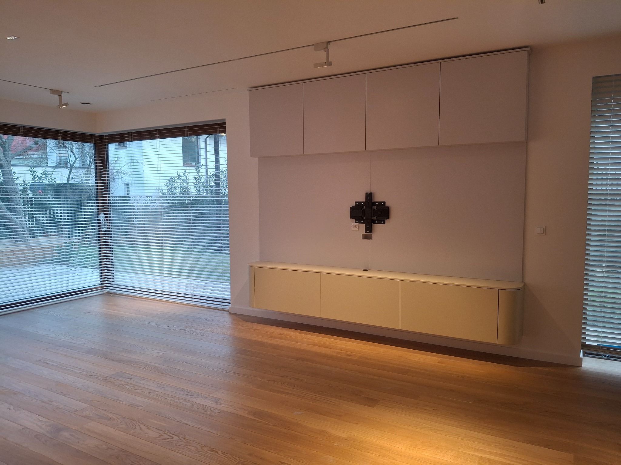 Jasne, minimalistyczne wnętrze z drewnianą podłogą, zabudową meblową na ścianie i dużymi oknami z żaluzjami. Widoczny uchwyt na telewizor.