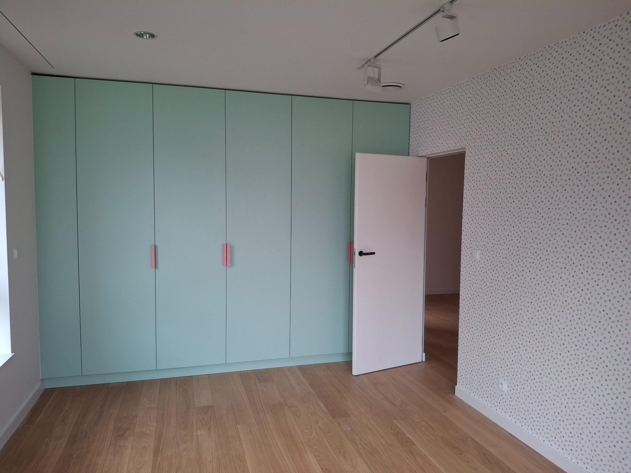 Pokój po remoncie: zabudowa szafy w kolorze miętowym z różowymi uchwytami, drewniana podłoga, ściana w tapetę w drobny wzór, białe drzwi otwarte.