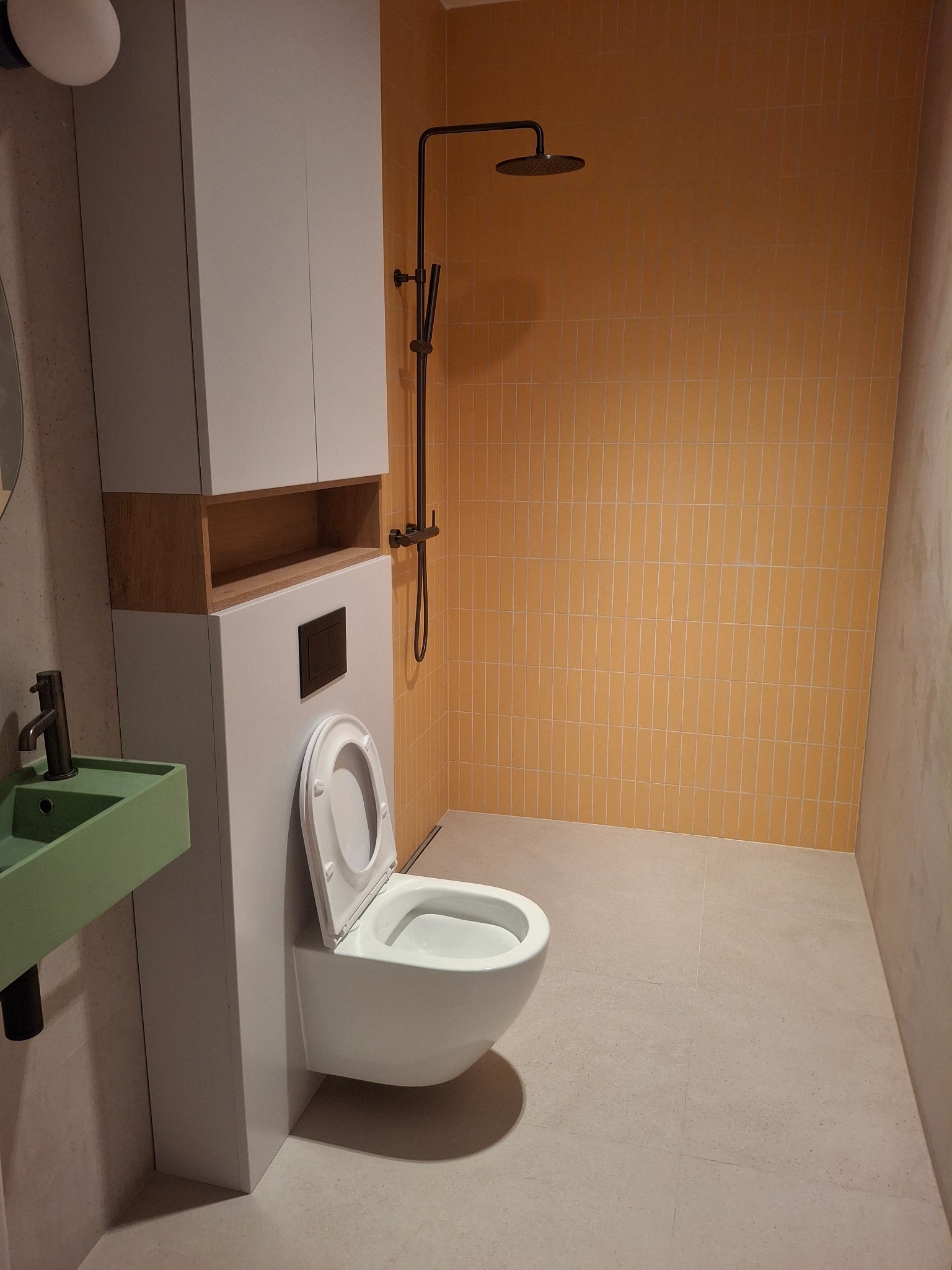 Nowoczesna łazienka z wiszącą toaletą, zieloną umywalką i pomarańczowymi płytkami pod prysznicem. Minimalistyczny design z elementami drewna.