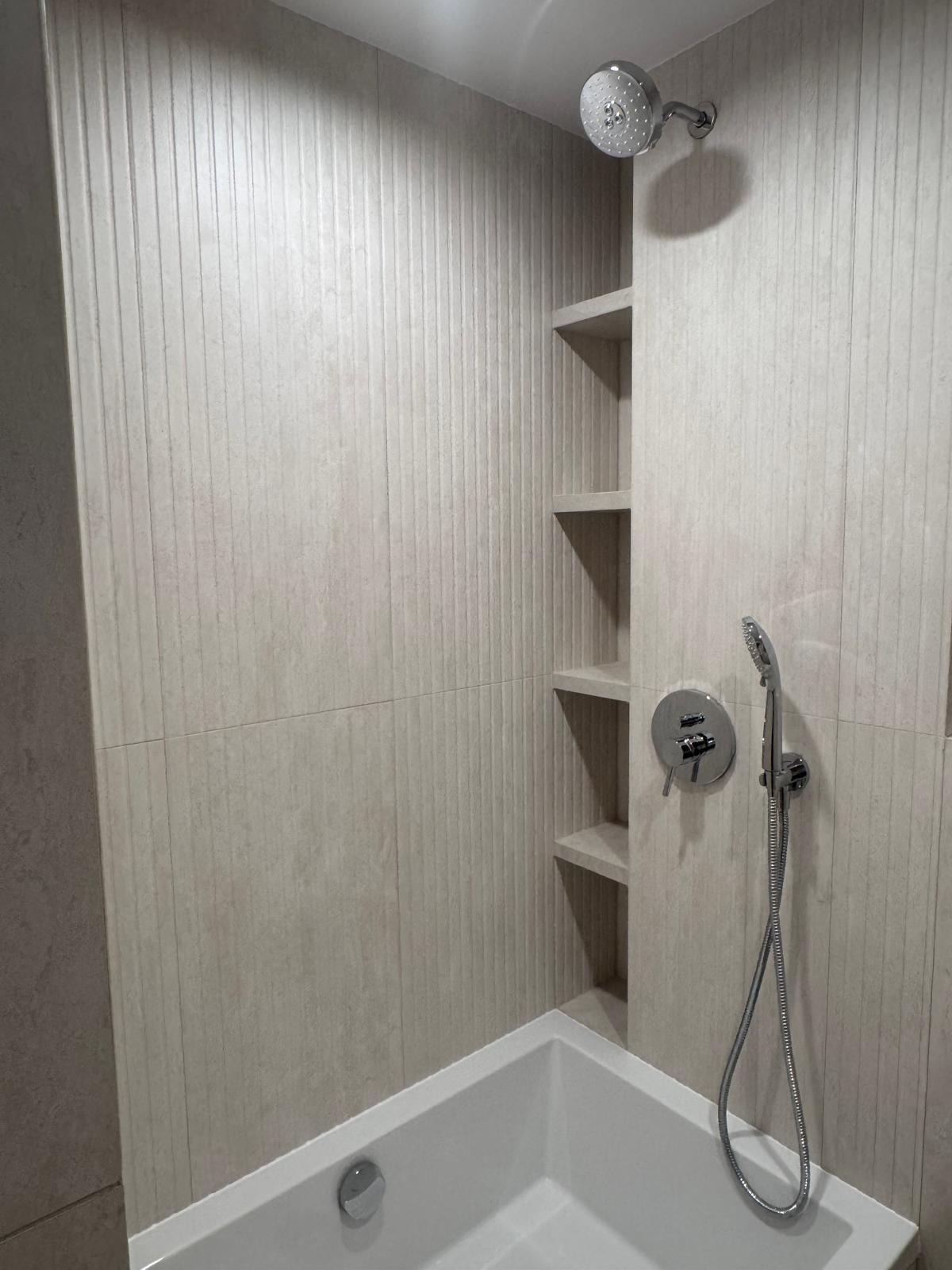 Wykończona łazienka z wanną, beżowymi płytkami z delikatnym wzorem pionowych pasków, wnękami pełniącymi funkcję półek i chromowaną armaturą.