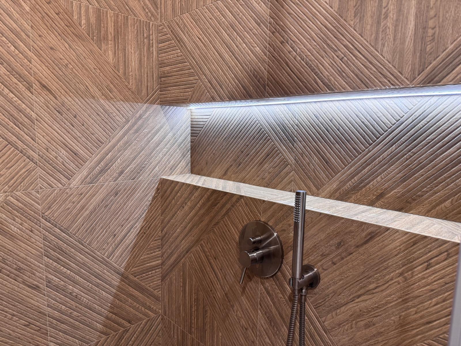 Nowoczesna kabina prysznicowa z drewnopodobnymi płytkami ułożonymi w geometryczny wzór. Bateria prysznicowa w kolorze stali nierdzewnej. Oświetlenie LED w półce.