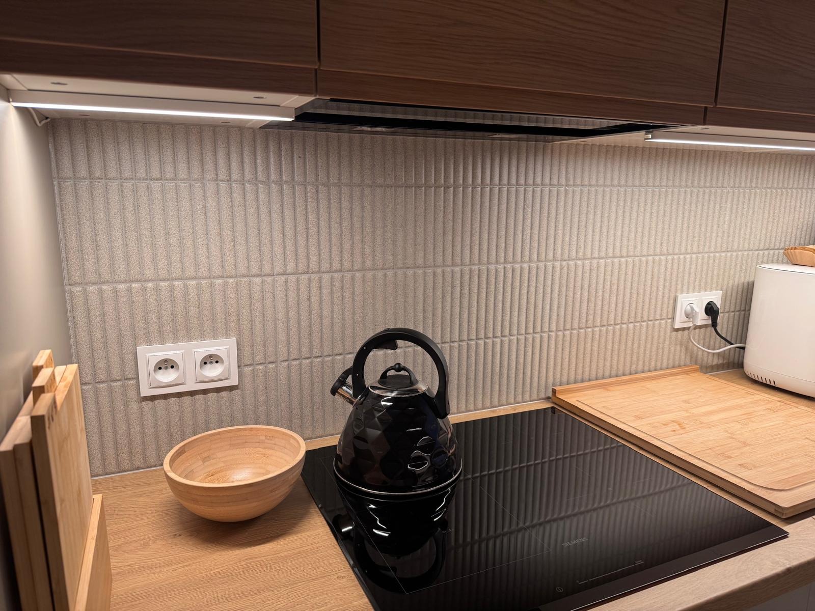 Beżowe, pionowe płytki na ścianie nad płytą indukcyjną w kuchni. Widoczny czarny czajnik, drewniana miska i deska do krojenia. Oświetlenie podszafkowe.