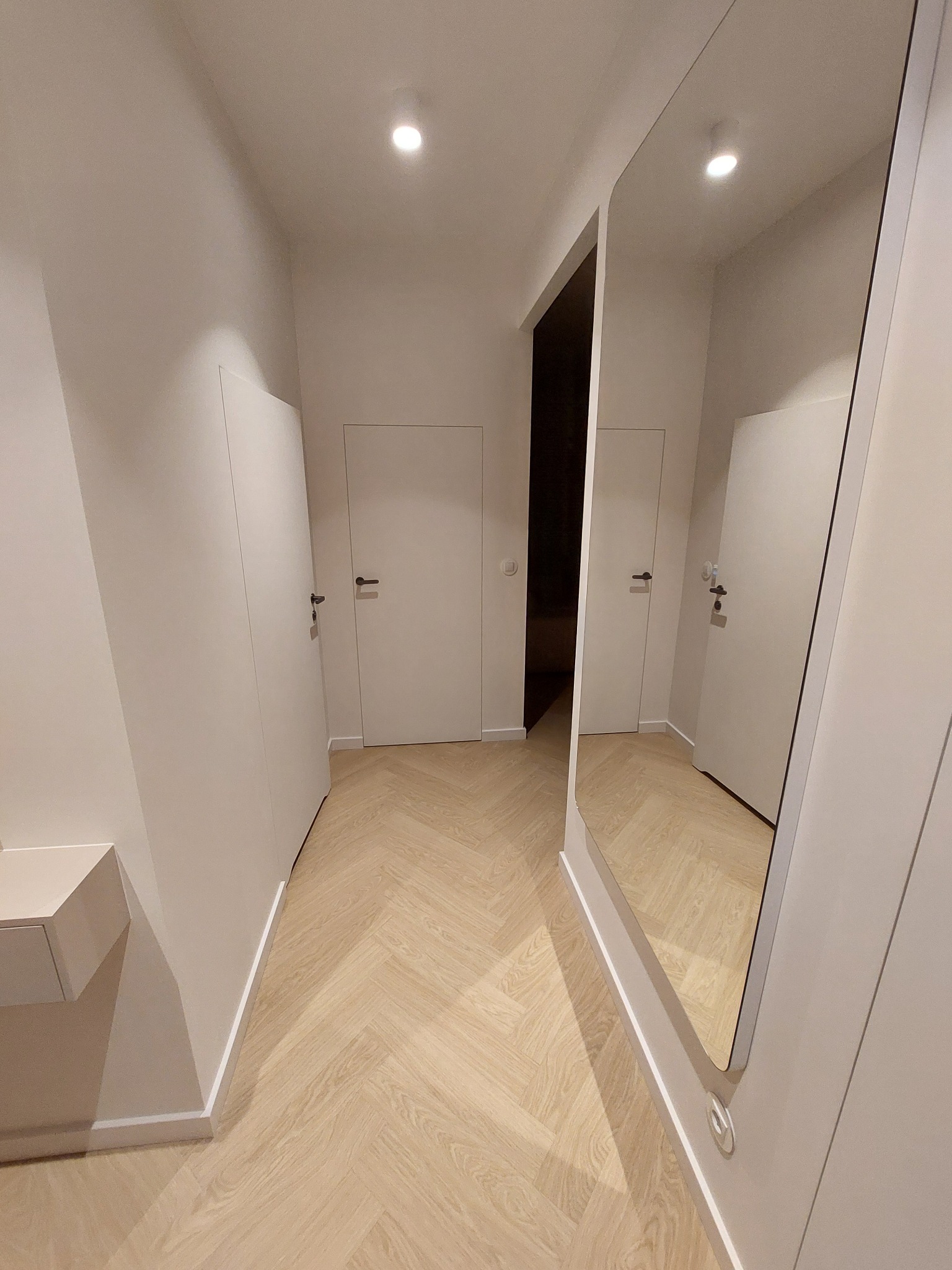 Jasny korytarz z parkietem w jodełkę, białymi drzwiami i dużym lustrem. Minimalistyczny design, subtelne oświetlenie punktowe. Widok na wnętrze mieszkania.