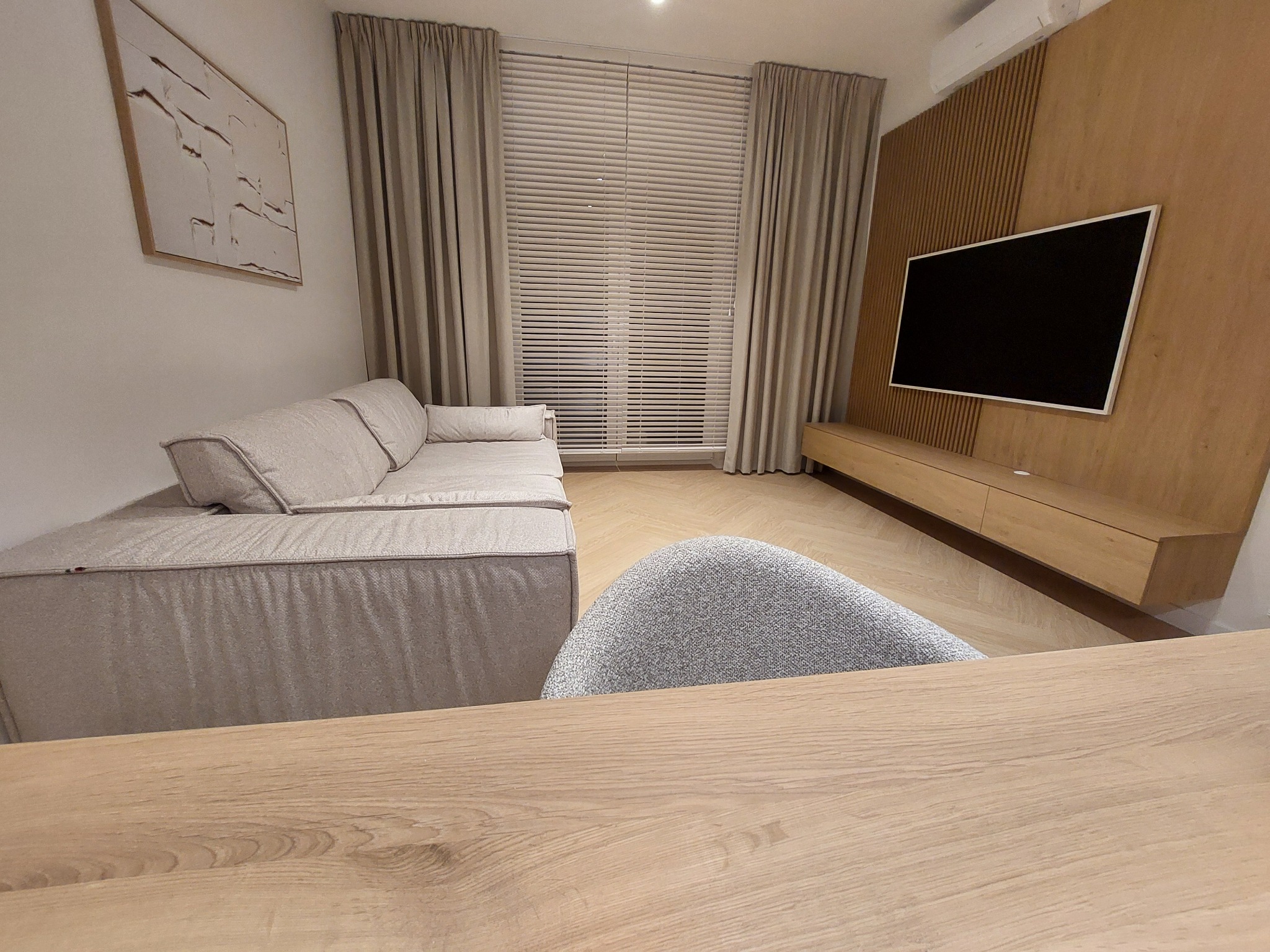 Jasne wnętrze salonu z beżową sofą, drewnianą ścianą z telewizorem i zasłoniętym oknem. Minimalistyczny design z elementami drewna i neutralną paletą barw.
