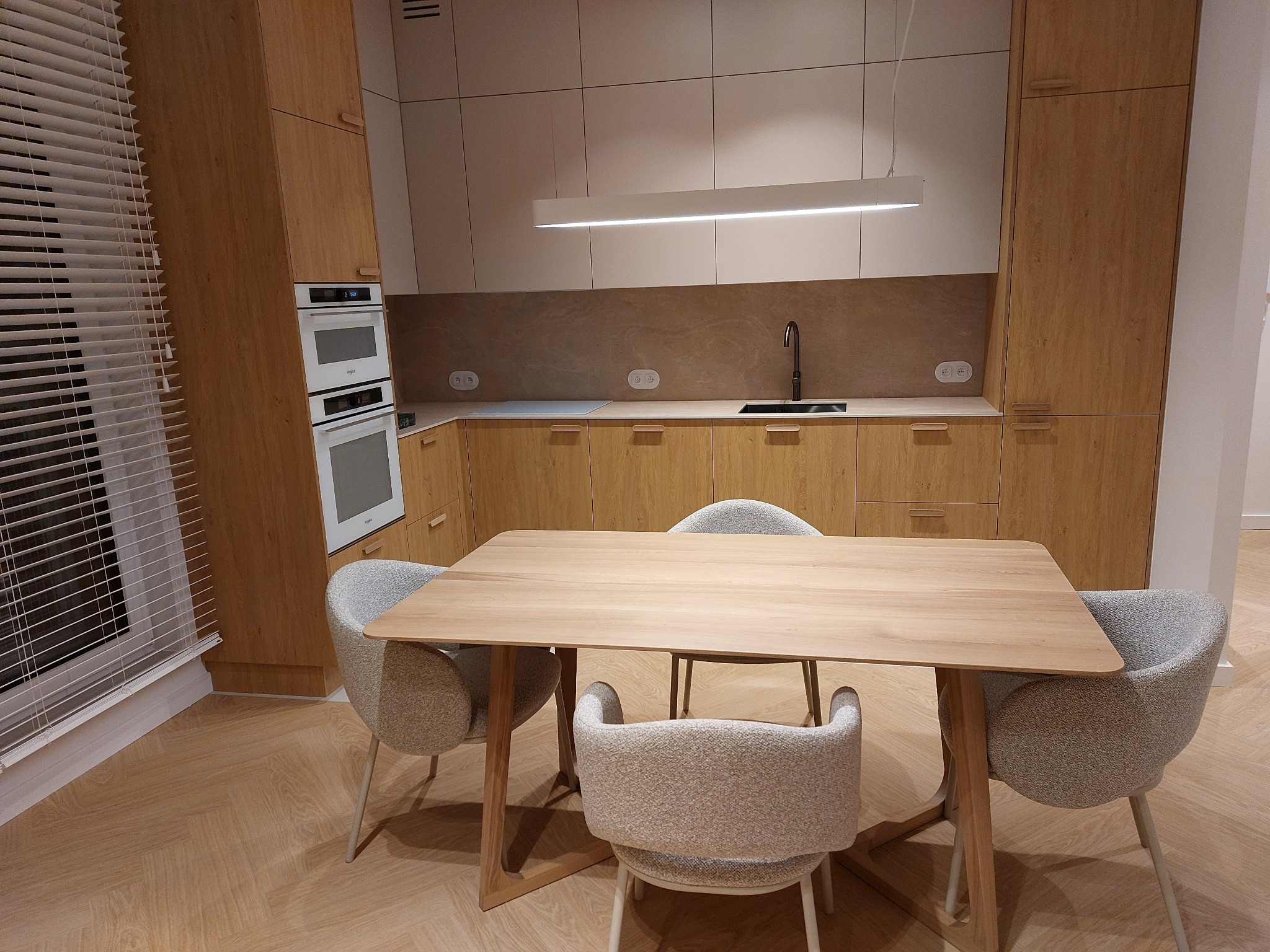 Jasna, nowoczesna kuchnia z drewnianymi frontami i stołem z krzesłami. Parkiet ułożony w jodełkę. Minimalistyczna lampa nad blatem. Okno z żaluzjami po lewej stronie.