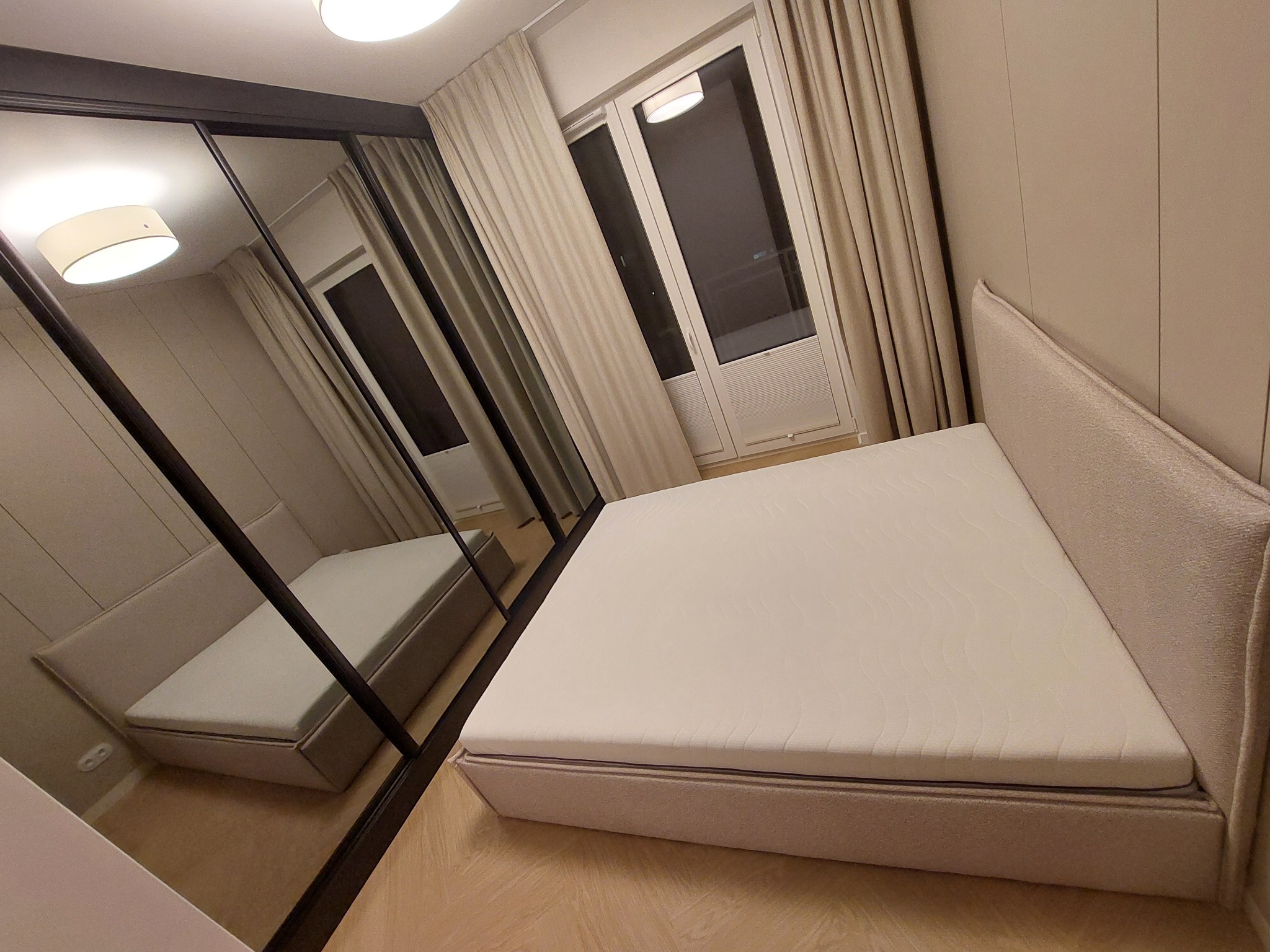 Wykończona sypialnia w neutralnych barwach w Warszawie. Widoczne łóżko z materacem i szafa z lustrzanymi drzwiami, odbijająca wnętrze pokoju.