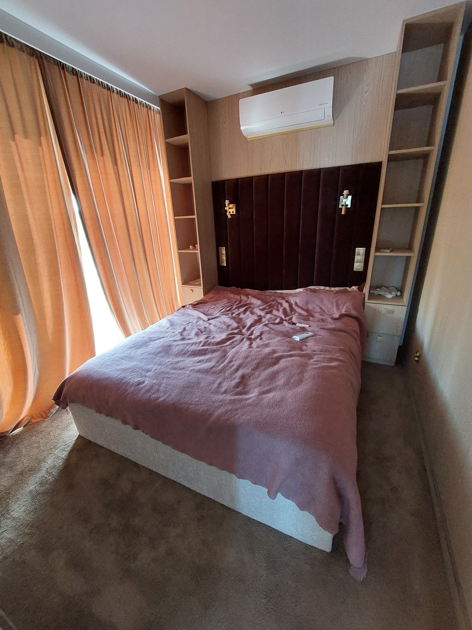 Nowoczesna sypialnia z brązowym zagłówkiem, drewnianymi półkami i beżowymi zasłonami. Klimatyzator na ścianie. Ustawienie ukośne, ukazujące przestrzeń i detale wykończenia.