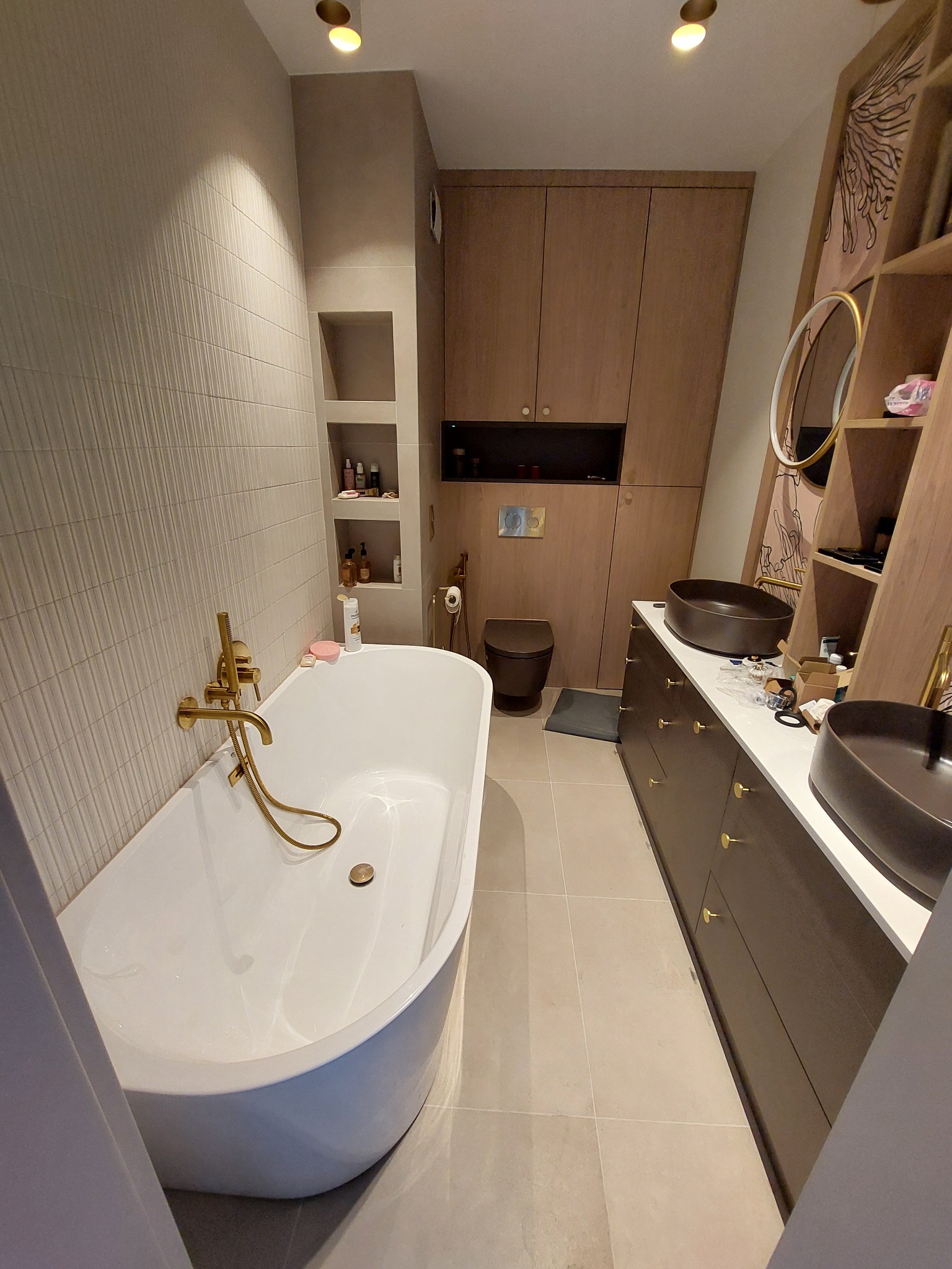 Nowoczesna łazienka z wolnostojącą wanną, złotą armaturą i umywalkami nablatowymi w odcieniach brązu. Minimalistyczne szafki i półki w zabudowie.