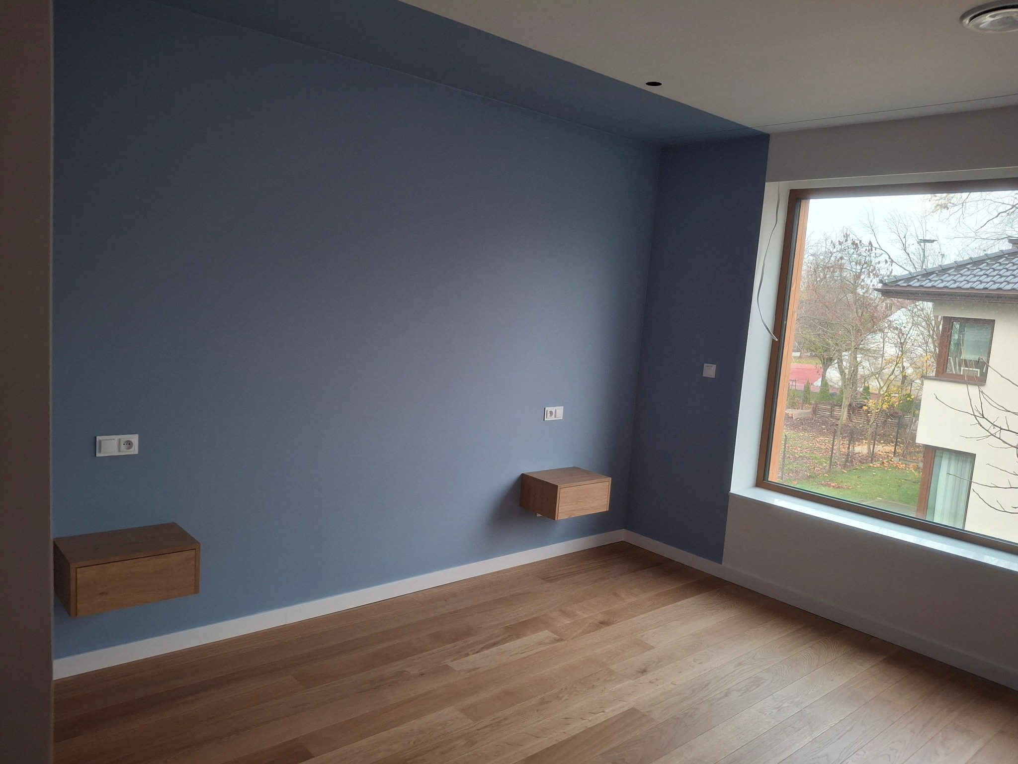 Pokój po remoncie: błękitne ściany, drewniana podłoga, okno z widokiem na zewnątrz, dwie wiszące szafki nocne i gniazdka elektryczne na ścianie.
