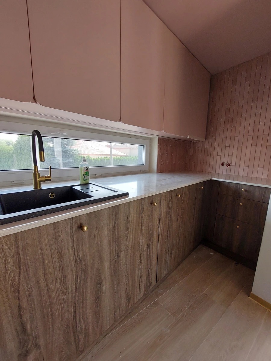 Nowoczesna kuchnia po remoncie: różowe szafki górne, drewnopodobne dolne, czarny zlew z złotą baterią, płytki na ścianie. Widok na zewnątrz przez okno.