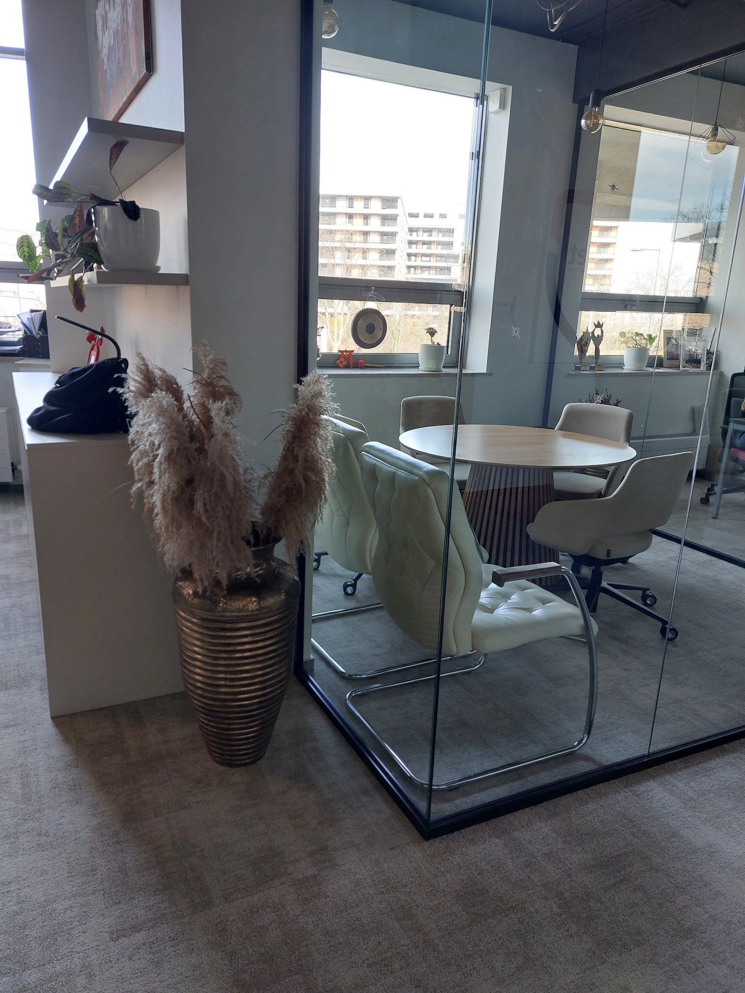 Nowoczesne biuro z przeszkloną salą konferencyjną, beżowe krzesła i okrągły stół. Wazon z pampasami dodaje przytulności. Widok na budynek za oknem.