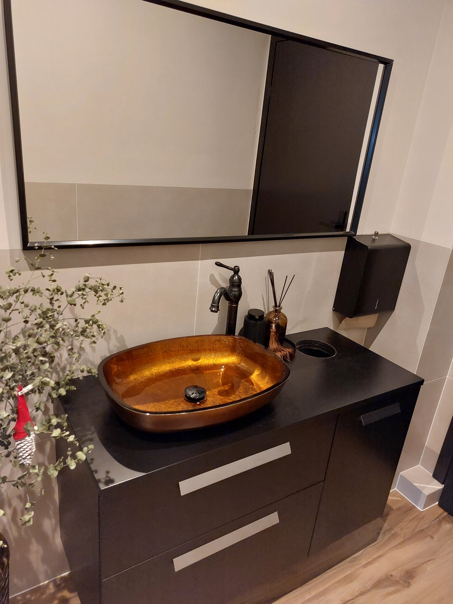 Nowoczesna łazienka z umywalką w odcieniach złota, czarną armaturą i szafką z szufladami. Minimalistyczne lustro i dekoracyjne gałązki dodają elegancji.