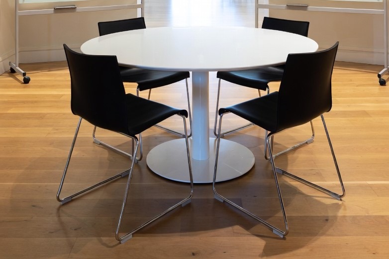 Biały okrągły stół z czarnymi krzesłami na parkiecie. Minimalistyczny design, jasne wnętrze, widoczne detale nóg krzeseł i struktury drewna podłogi.