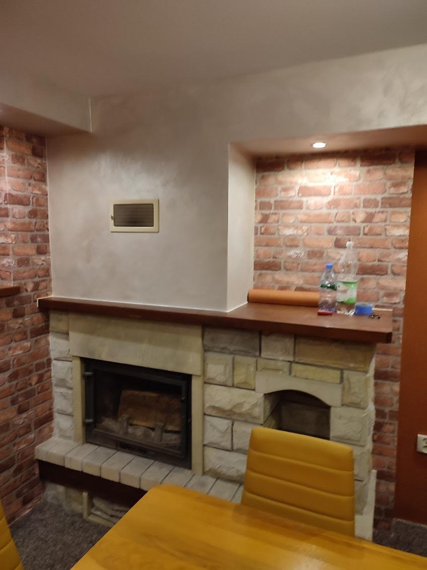 Kamienny kominek z drewnianym blatem, otoczony ścianą z cegły i gładzią. Na blacie butelki z wodą i zwinięty materiał. Część stołu i krzesła w kadrze.